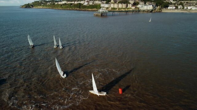A tilting up drone shot of small sailing boats rounding a buoy the shot then tilts up to reveal Clevedon Pier and Coastline