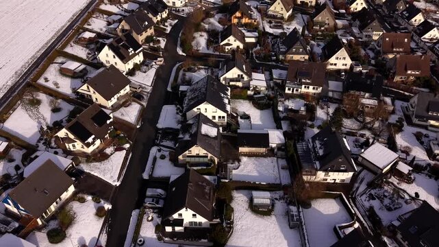 Aerial flyover snow-covered single family houses in German suburban neighborhood, showing quiet residential streets, winter conditions, rooftops, gardens and calm suburban environment. Top down shot.