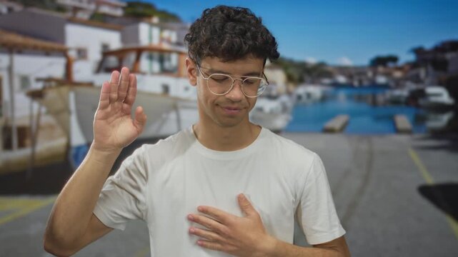 Man raises hand to wave by a building and smiles softly while the other hand rests on chest; sincerity.