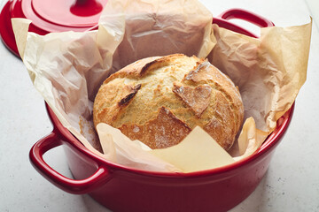 Golden sourdough bread with cracked crust in red ceramic Dutch oven, lined with parchment paper. Perfect for culinary, baking, and food photography projects.