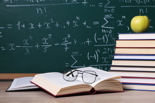 Open book with eyeglasses and tall stack of textbooks, green chalkboard covered with math equations and formulas in background

