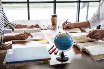Two people studying together at table with books, globe, coffee and sticky notes, collaborative learning and tutoring in bright room


