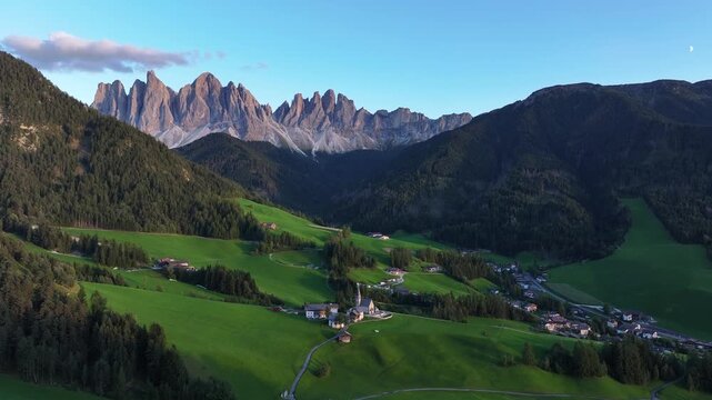 Aerial view of the Santa Magdalena church nestled among vibrant green fields and towering, jagged mountains, Santa Magdalena, Trentino-Alto Adige, Italy.