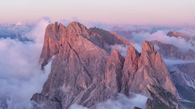 Aerial view of jagged Seceda mountain peaks piercing through a sea of clouds, their rocky faces illuminated by the soft glow of dawn, Seceda, Trentino-South Tyrol, Italy.