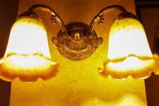 Warm vintage wall sconce casting glow. Ornate brass fixture with frosted glass shades mounted on textured plaster wall, bathing corridor in amber light closeup reveals filigree details, polished