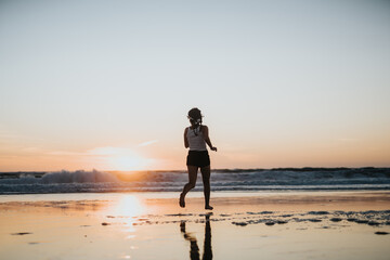 Fototapeta premium A silhouette of a woman jogging on wet sand during sunset, with waves rolling in the distance. Warm light creates reflections, conveying fitness, freedom, and outdoor serenity.