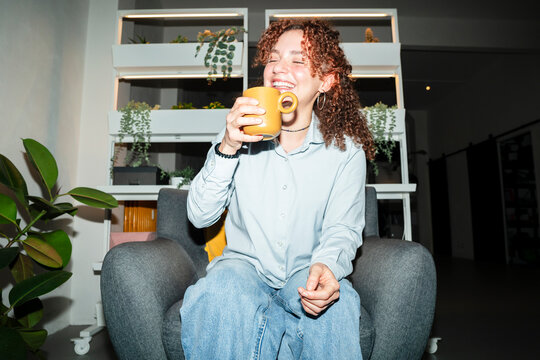 A young woman with curly hair laughs while drinking from a mug, seated comfortably in an armchair in a modern living space.