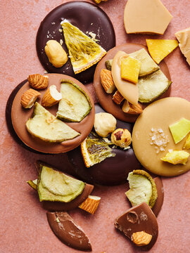 Gourmet dark and milk chocolate discs with various dried fruits like apple, orange, and mango, plus whole and chopped nuts, arranged on a textured pink background