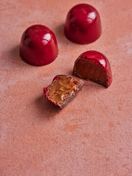 Three bright red dome chocolates rest on a textured pink surface. One is sliced to reveal a luscious, golden-brown caramel filling. Two chocolates remain whole, showcasing their shiny, vibrant shells