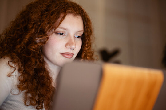 A young woman with curly red hair looks intently at a tablet screen, her face illuminated by its glow.