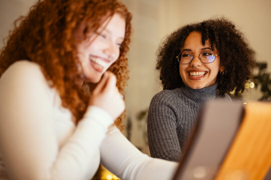 Two young women share a moment of laughter while looking at a screen, their faces illuminated by its glow.