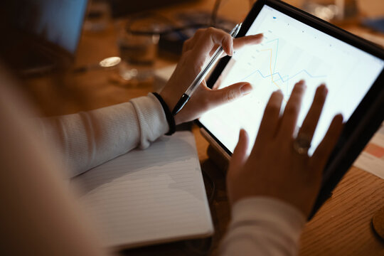 A person's hands interact with a tablet displaying financial charts and data, with a notebook and pen nearby, suggesting analysis and planning.