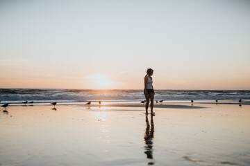 A woman in casual beachwear stands on the wet sand as the sun sets over the ocean. Seabirds dot the shore, creating a peaceful, reflective moment by the water. © qunica.com