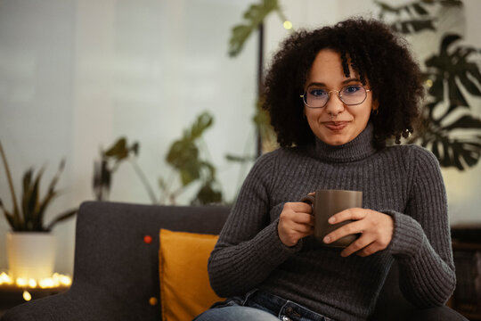 A young woman with curly hair and glasses smiles while holding a mug, seated in a cozy armchair with plants in the background.