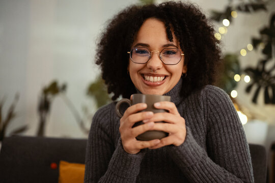 A young woman with curly hair and glasses smiles while holding a mug of a warm beverage.