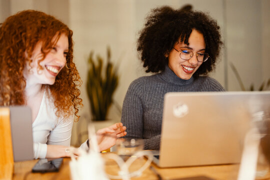 Two young women with curly hair are smiling and working together on a laptop in a casual setting.
