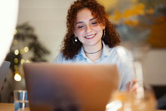 A young woman with curly hair smiles while working on her laptop in a softly lit room with bokeh lights in the background.