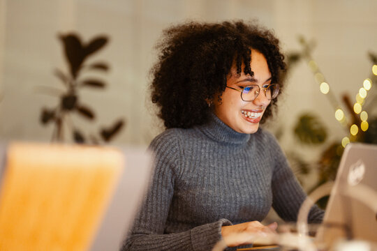 A young woman with curly hair and glasses smiles while working on a laptop in a cozy, softly lit room with plants and string lights.