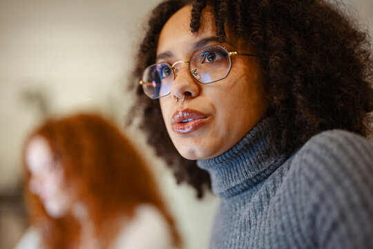 A young woman with curly hair and glasses speaks thoughtfully, her gaze directed upwards. Another person is out of focus in the background.