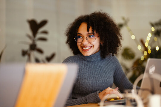 A young woman with curly hair and glasses smiles while working on her laptop in a cozy, softly lit environment.