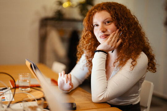 A young woman with curly red hair uses a tablet at a wooden table, holding a pen and looking towards the camera.