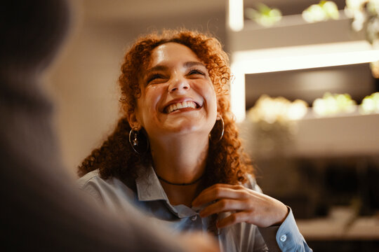 A young woman with curly hair laughs heartily while looking up at someone. Her eyes are closed in joy, and her hand is raised to her chest.