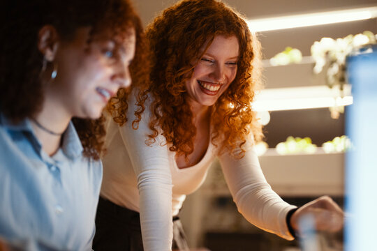 Two young women with curly red hair share a moment of laughter while working together on a project, illuminated by warm indoor lighting.