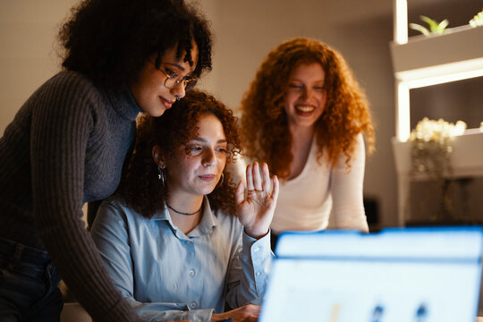 Three young women collaborate on a project, one raising her hand as if to speak during a video call.