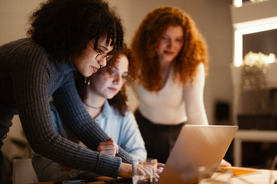 Three young women collaborate on a project, gathered around a laptop. One woman leans in, pointing at the screen, while another looks on intently.