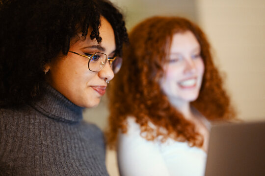 Two young women with curly hair are looking at a screen, one wearing glasses and a grey turtleneck, the other smiling in the background.