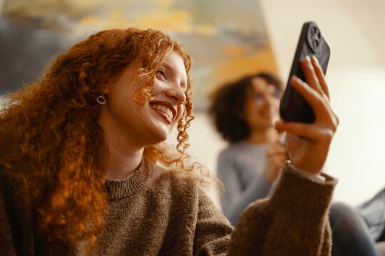 A young woman with curly red hair smiles as she holds up her phone, capturing a moment with a friend in the background.