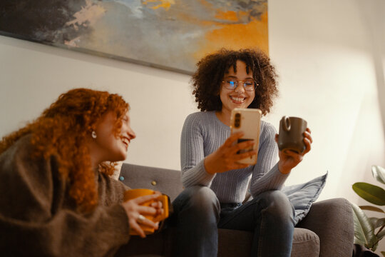 Two young women share a laugh while relaxing on a sofa with drinks and a mobile device.