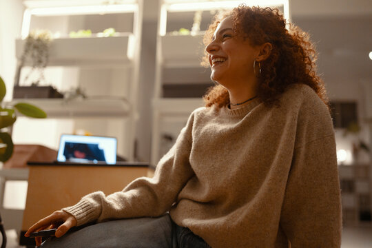 A young woman with curly hair laughs while holding a remote control, relaxing in a cozy room with a laptop in the background.