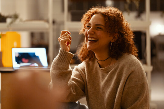 A young woman with curly red hair laughs heartily while sitting in a dimly lit room with a laptop open in front of her.
