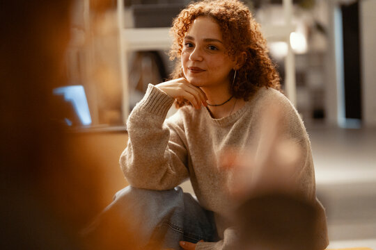 A young woman with curly red hair sits indoors, resting her chin on her hand, looking thoughtfully towards the viewer.