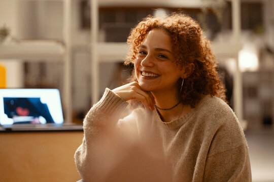 A young woman with curly red hair smiles while resting her chin on her hand, with a laptop open in the background.