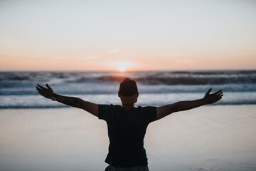 A man stands on a calm beach at sunset, arms wide open, conveying freedom, peace, and relaxation as the waves roll in and the sky glows with warm colors. © qunica.com