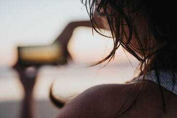 A woman stands on the beach at sunset, back to the camera, capturing the horizon with a smartphone. Warm light, flowing hair, and a calm, candid mood. © qunica.com