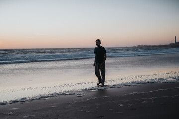 Naklejka premium A solitary man strolls the shoreline as gentle waves roll in during sunset, evoking calm, contemplation, and the serene mood of coastal life.