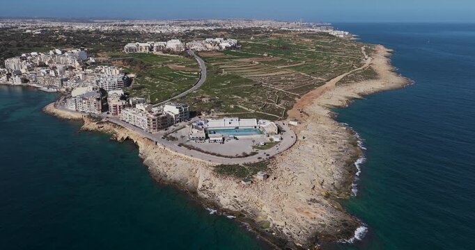 Aerial view of the rugged coastline meeting the buildings of Marsaskala, with the turquoise sea contrasting against the rocky shore, Marsaskala, Wied il-Ghajn, Malta.
