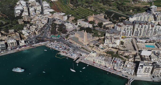 Aerial view of the Marsaskala parish church and harbor where boats rest on the tranquil water, contrasting with the surrounding buildings, Marsaskala, Wied il-Ghajn, Malta.
