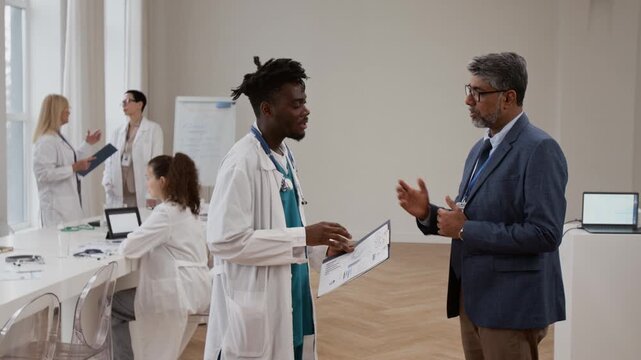Multiethnic mature formally dressed doctor talking with Black young colleague in lab coat, using thumps up gesture to show his approval and shaking hands with man during medical convention