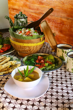 various traditional Indonesian dishes, including rice in a bamboo basket, fried chicken, assorted vegetables, and fresh drinks.