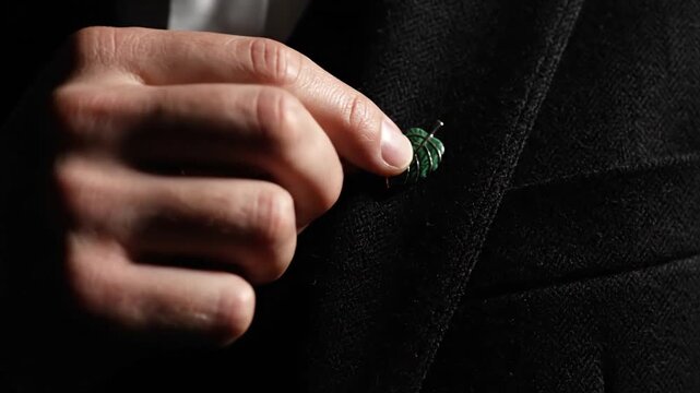 The video captures a hand meticulously fastening a delicate, carved green leaf lapel pin onto a dark, textured suit jacket, highlighting precision and elegance in formal dressing.