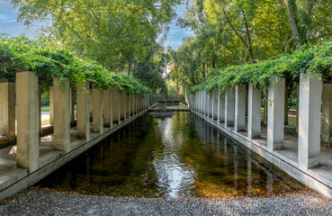 Fototapeta premium Paris, France - 08 15 2025: Bercy Park. Panoramic view of two covered and shaded walkways with concrete pillars and a basin between the two
