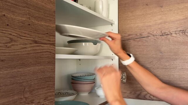 Placing clean dishes into kitchen cabinet after washing. Home organization routine and everyday kitchen maintenance concept.