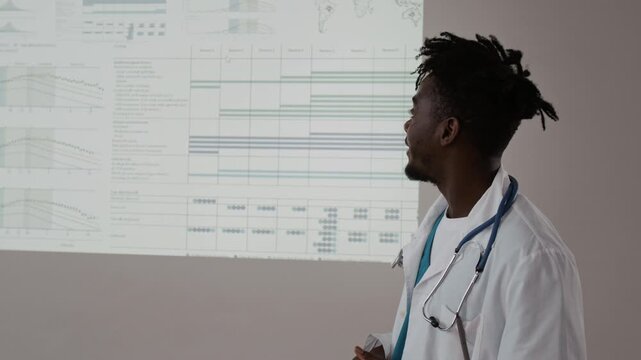 Black young male doctor in lab coat with stethoscope around his neck looking at interactive wall and referring to unknown audience while giving presentation during medical convention