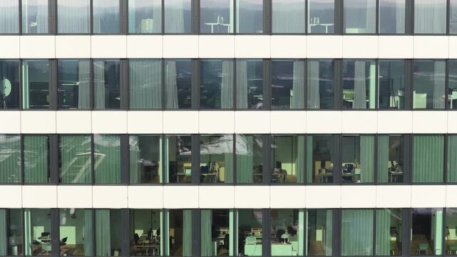 Aerial shot moving up along facade of a modern office building revealing employees working inside through glass windows