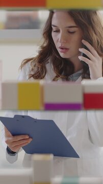 Female Pharmacist Talking on Smartphone Checking Medicines with Clipboard