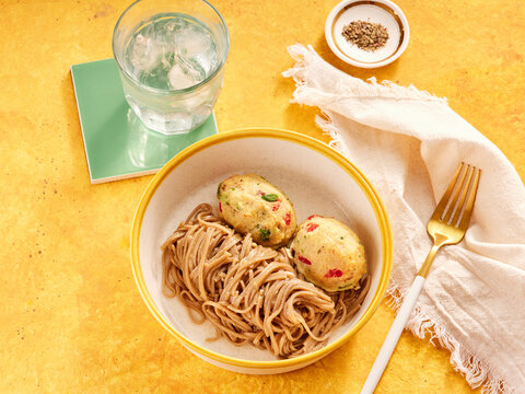 A white bowl holds soba noodles and two vegetable dumplings on a bright yellow surface